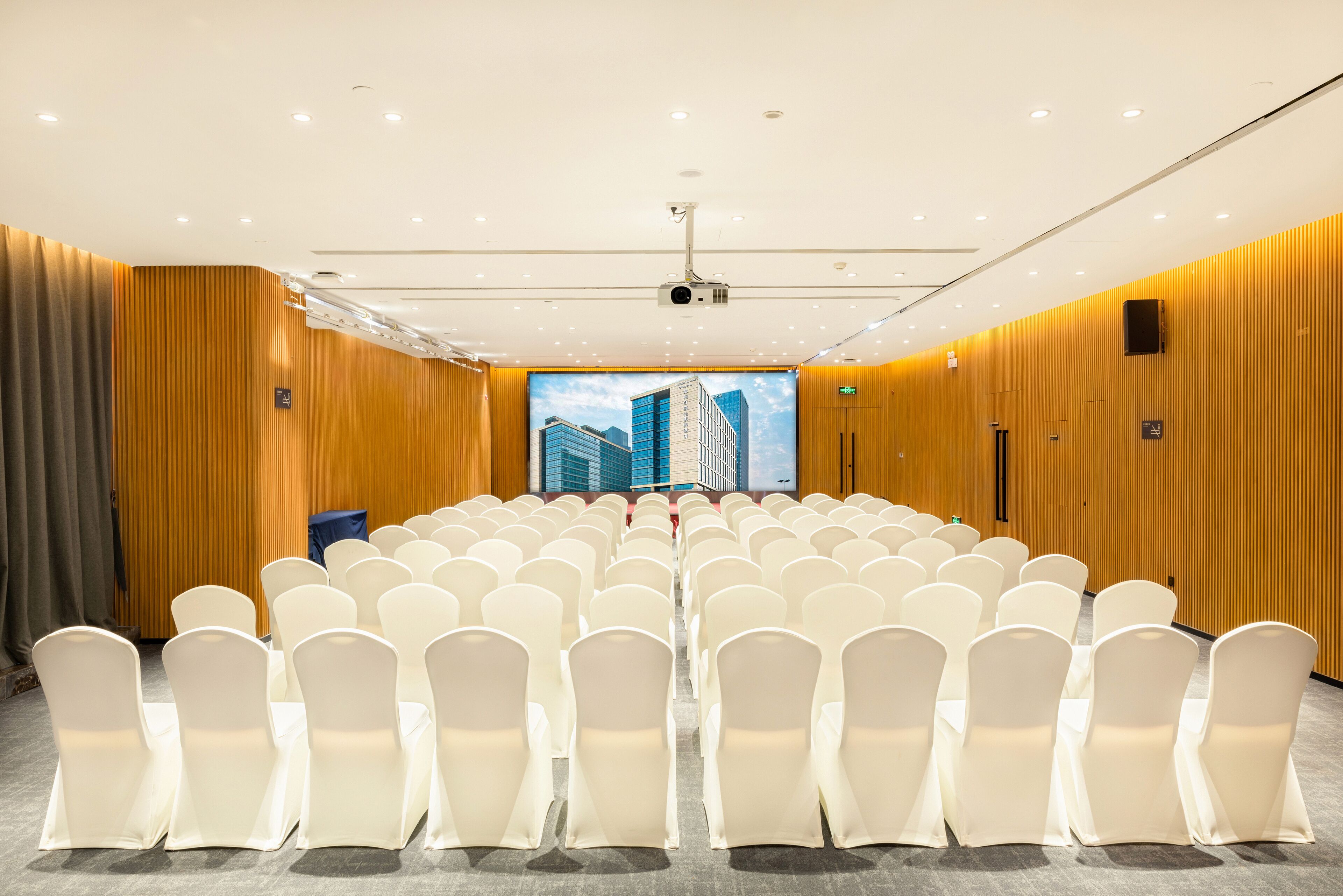 A conference room with rows of white chairs, a large screen at the front, and wooden paneling on the walls.