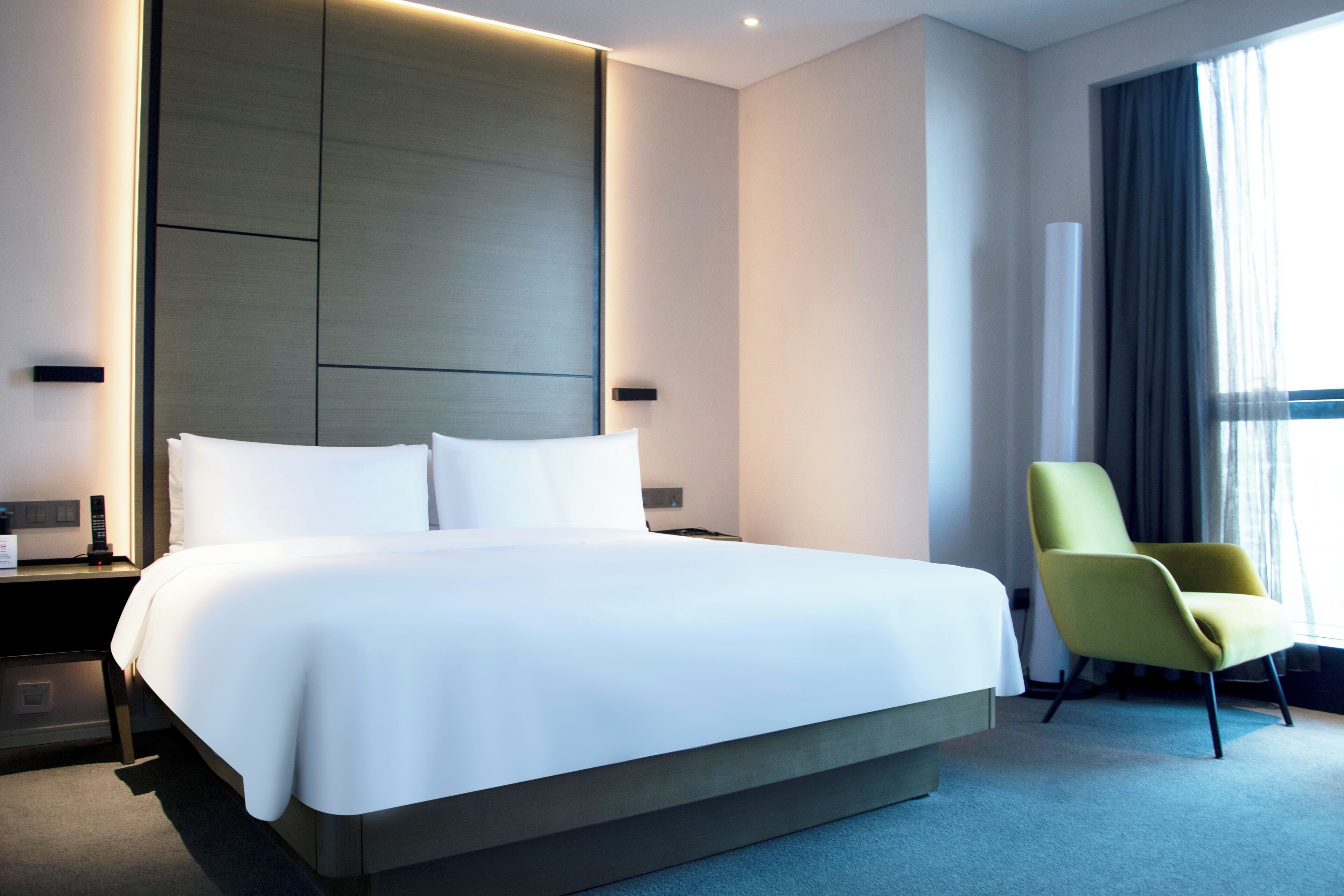 Una habitación de hotel moderna con una cama grande, una mesita de noche, una silla y un ventanal con cortinas.