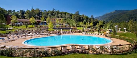 A large outdoor swimming pool surrounded by lounge chairs and a wooden fence, with a backdrop of buildings and hills.