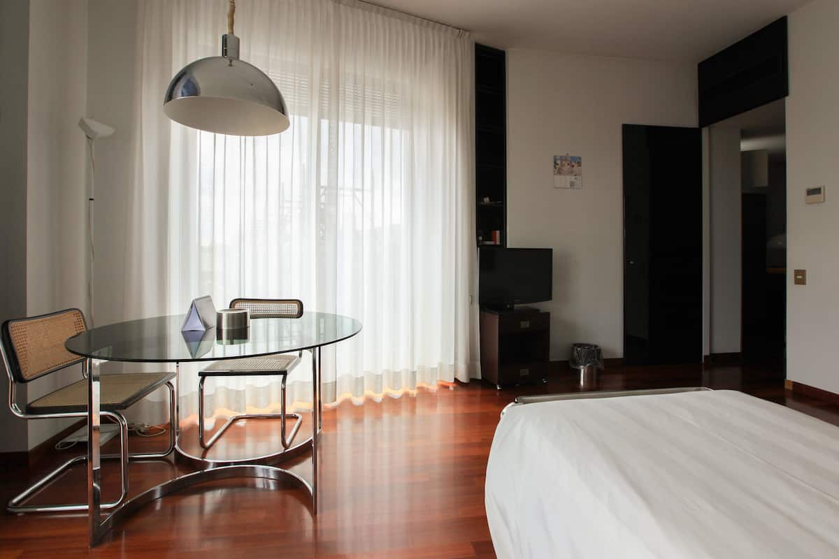 Habitación de hotel con cama, una mesita de vidrio con sillas, televisor y ventana con cortinas translúcidas.