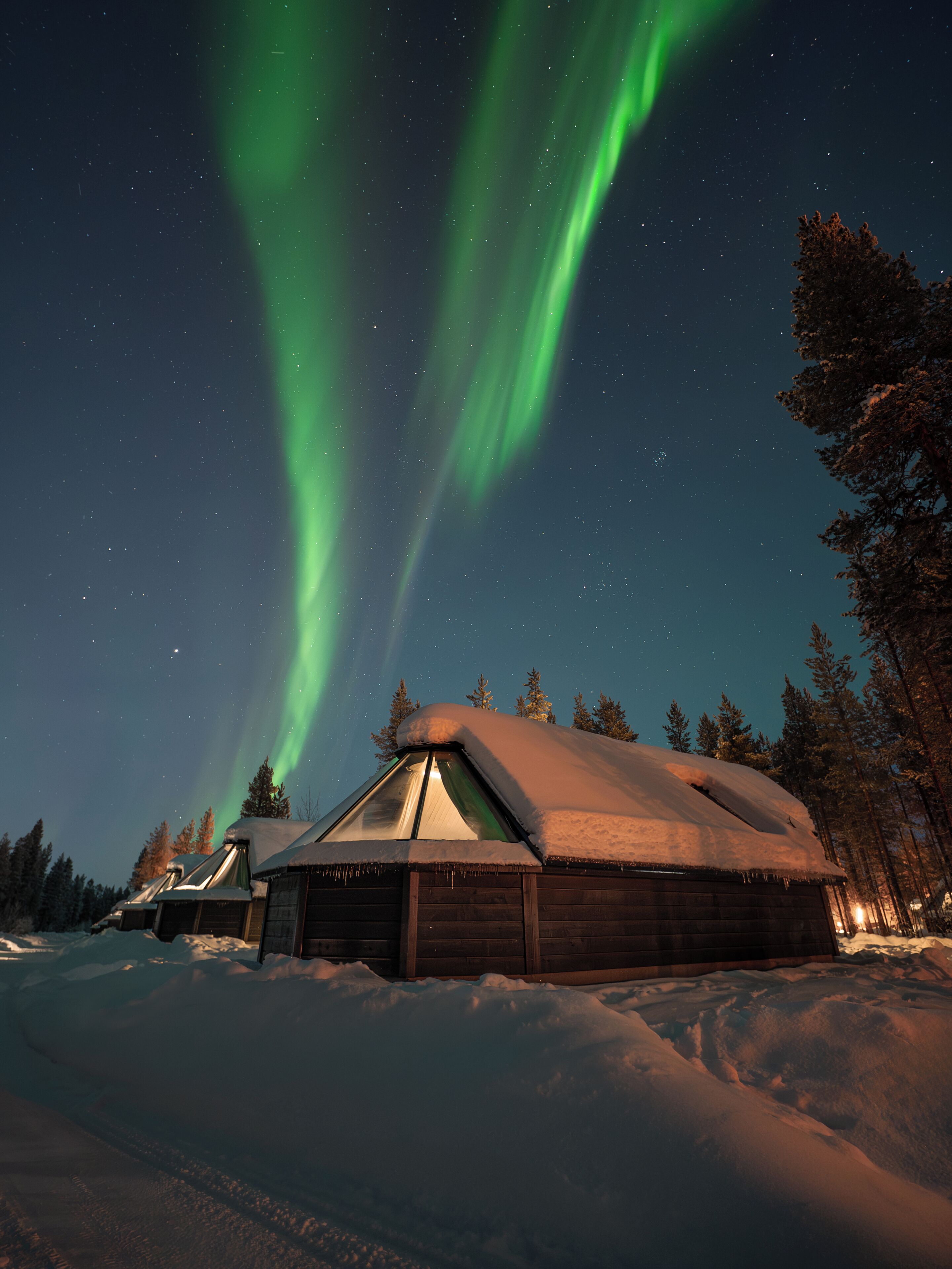 A cabin with a snow-covered roof, surrounded by trees, under a night sky with visible stars and green aurora.