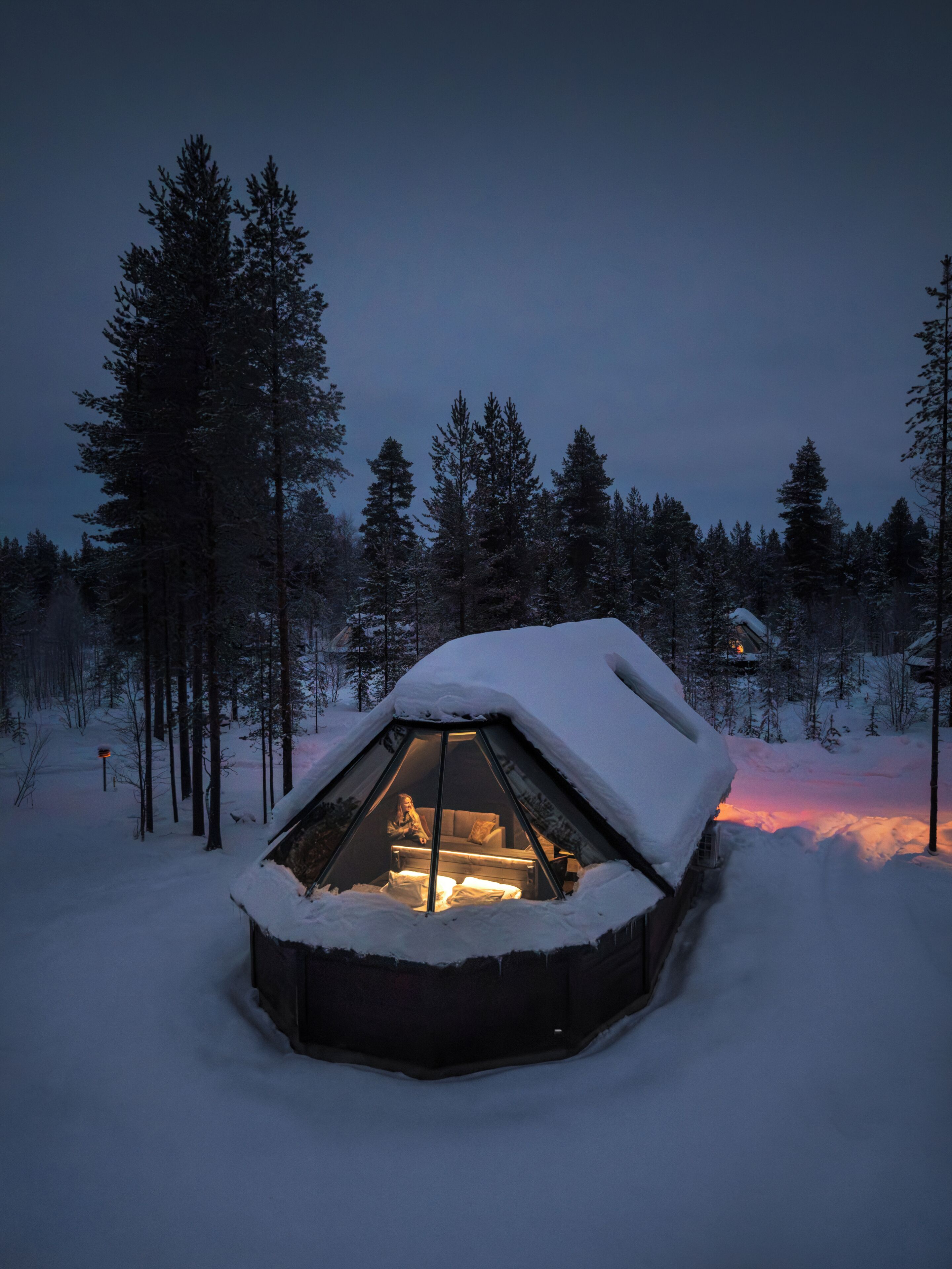 A snow-covered, hexagonal-shaped cabin with a lit interior, surrounded by a forest.