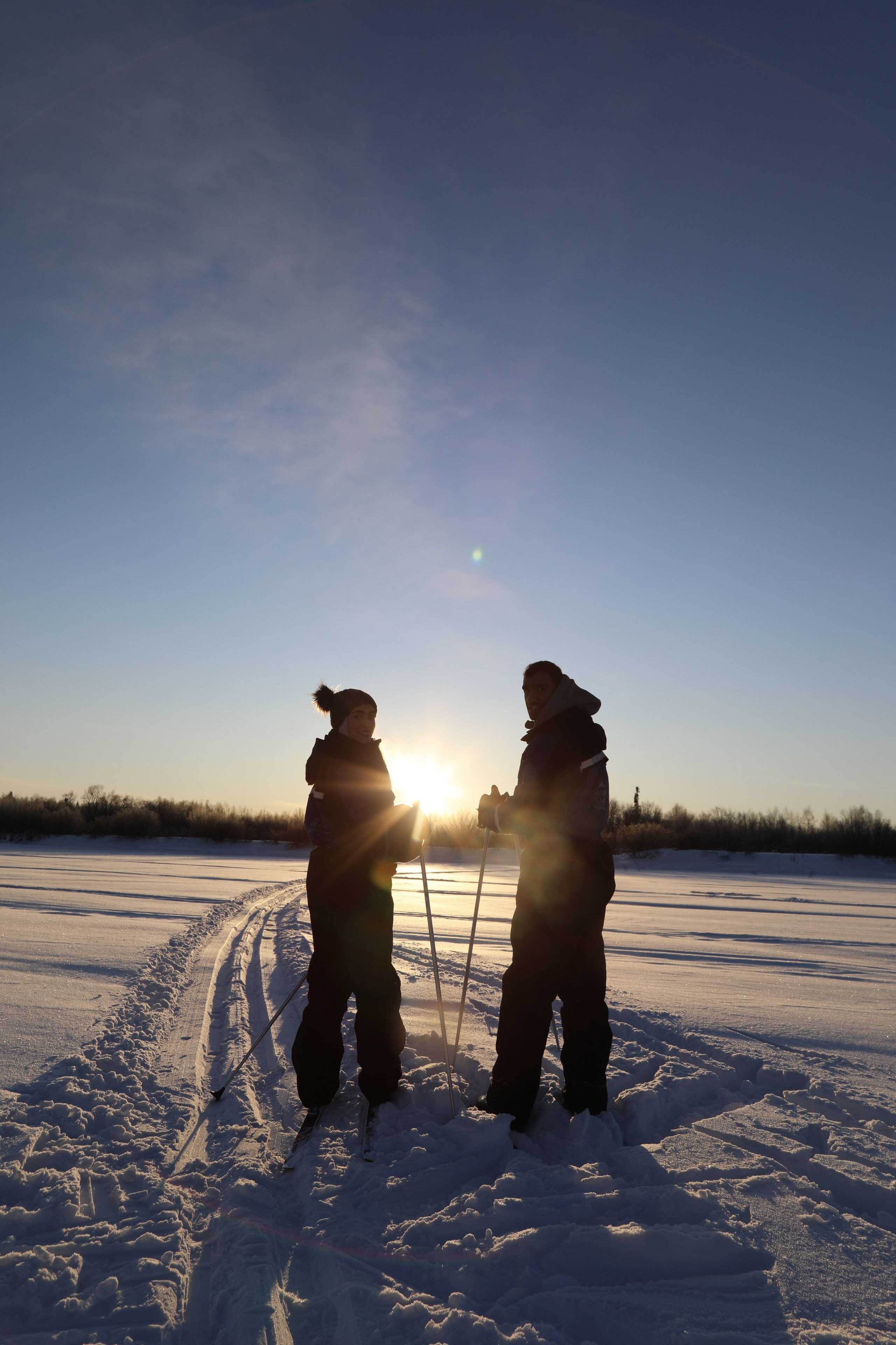 Duas pessoas esquiando em uma trilha nevada, com o sol se pondo ao fundo.