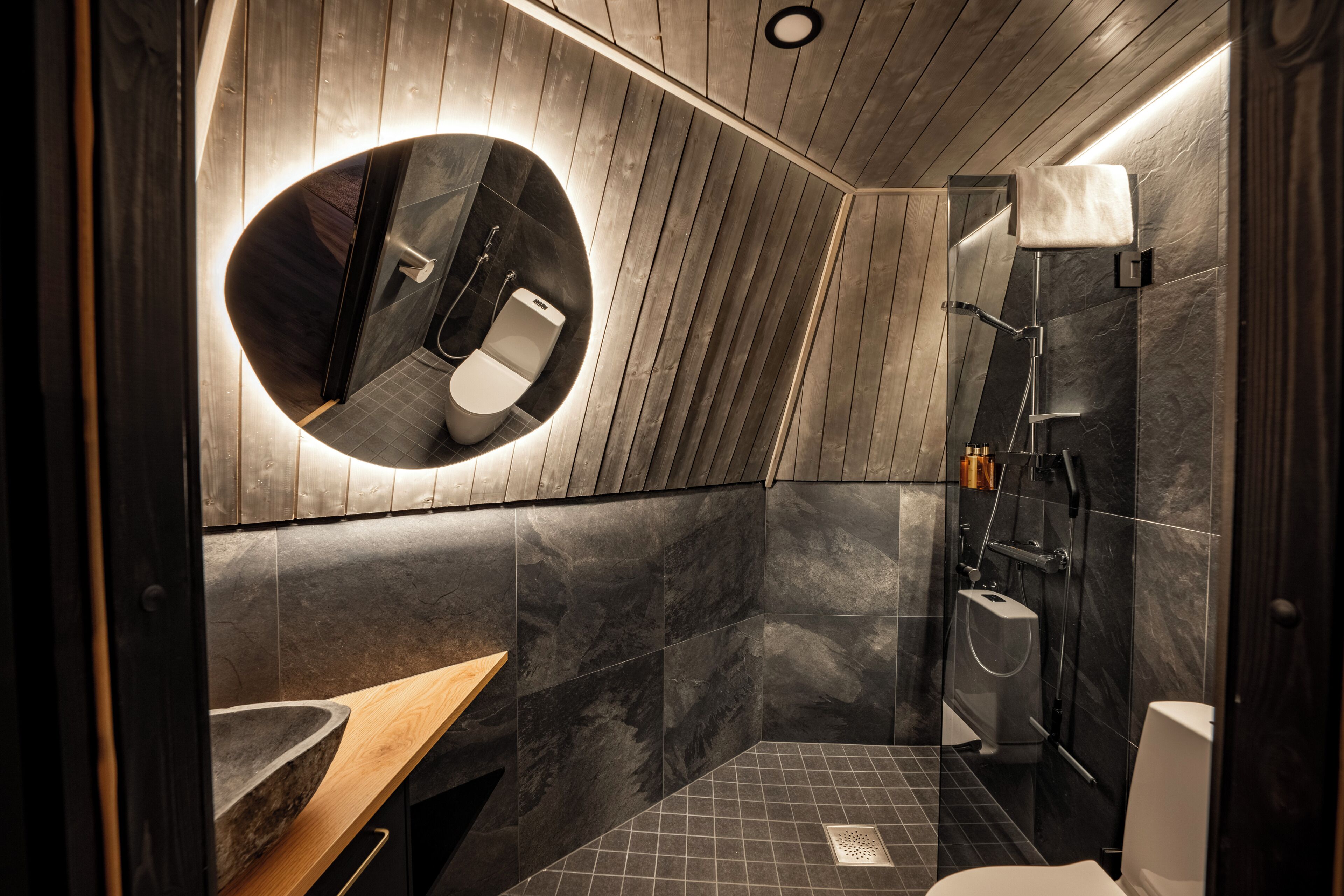 Un baño moderno con techo de madera, un espejo redondo, un lavamanos y una zona de ducha.