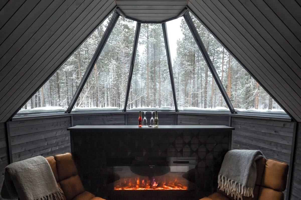A cozy room with a fireplace, two leather chairs, a round table, and a view of a snowy forest through large windows.