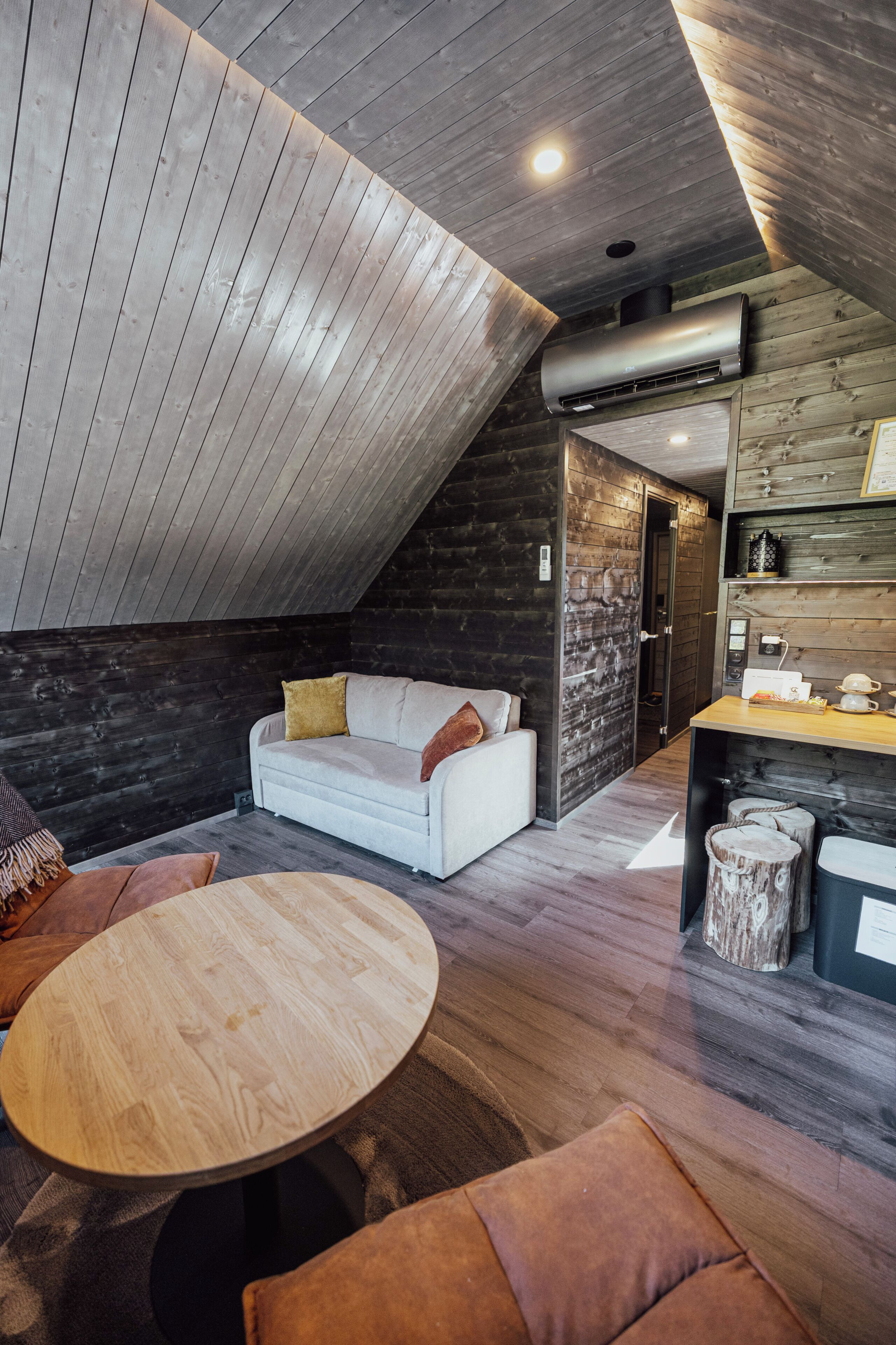 Un espai tipus loft amb sostre inclinat, un sofà blanc, una taula de fusta i una zona de cuina amb fogó i prestatges.