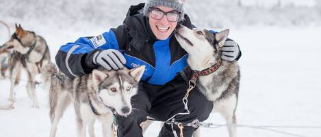Una persona con chaqueta azul y pantalones negros interactúa con un perro husky en un entorno nevado.