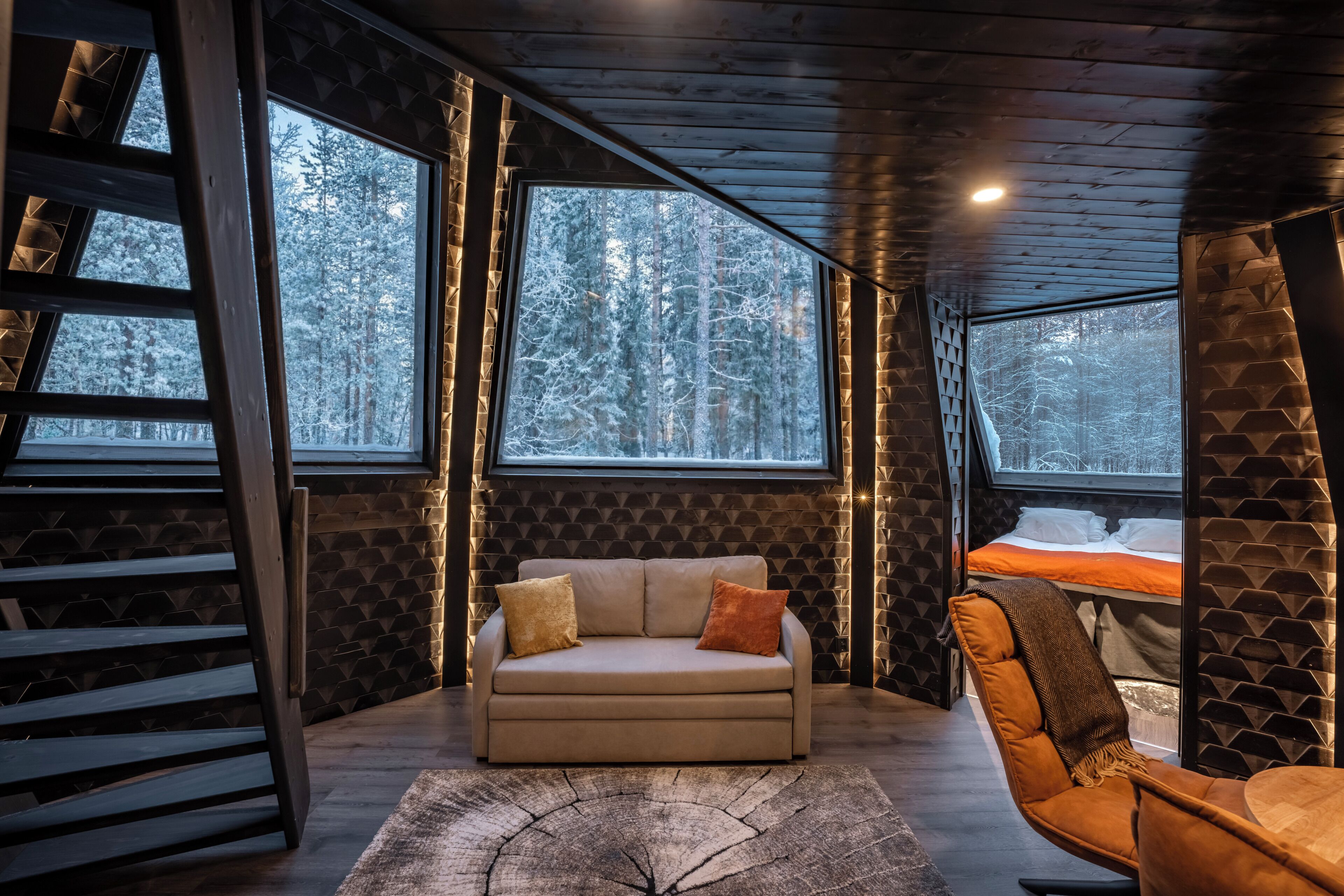 A cozy room with a sofa, a chair, a wooden floor, and large windows with a snowy forest view.