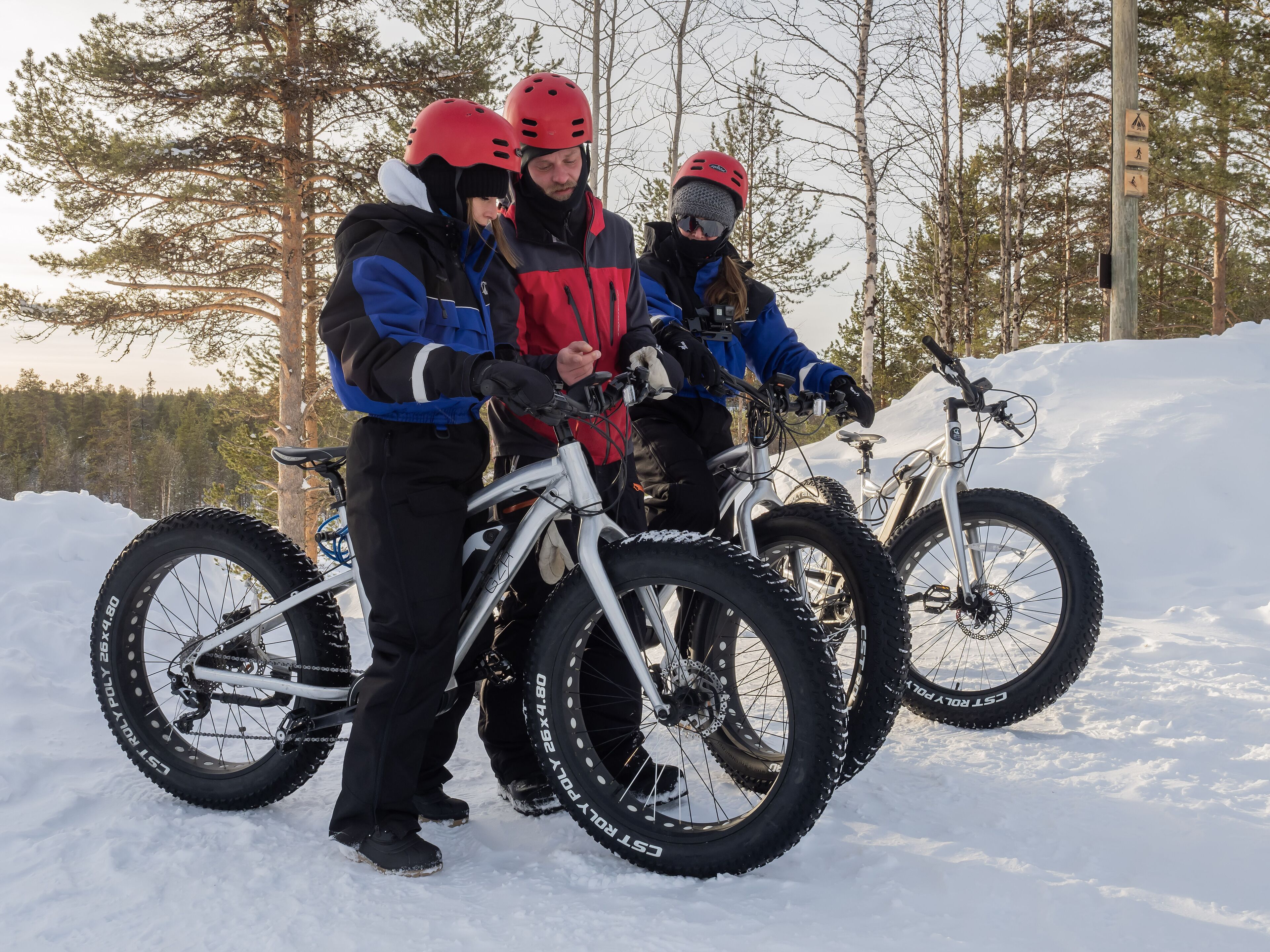 Three people standing next to bicycles in a snowy area with trees in the background.