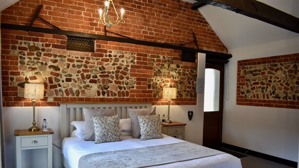 A bedroom with a brick wall, a bed with white bedding, two bedside tables with lamps, and a chandelier. - At the Farmhouse Hunters Hall (Dereham)