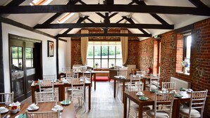 A dining area with multiple tables and chairs, large windows with curtains, and a brick wall. - At the Farmhouse Hunters Hall (Dereham)