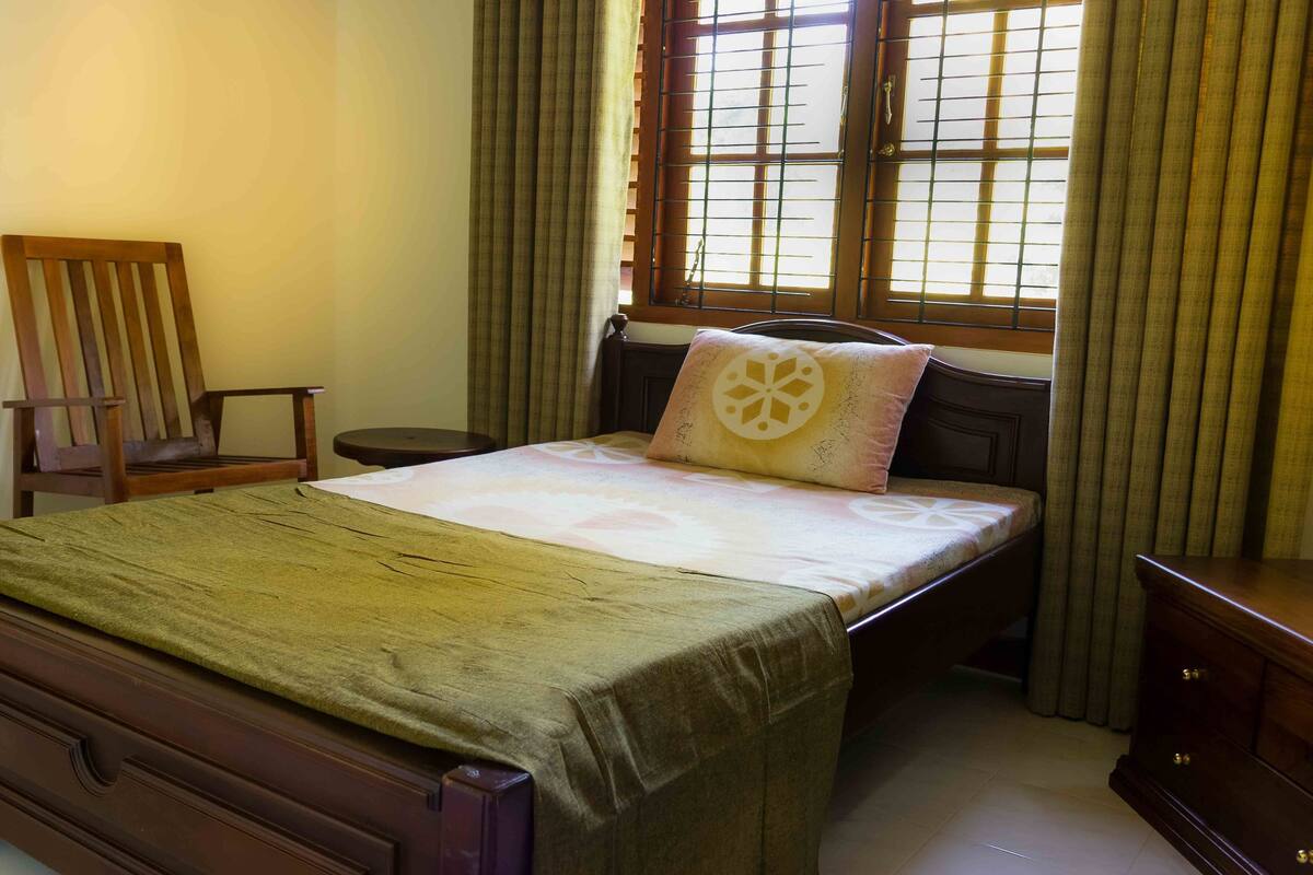 A bedroom with a bed, a wooden chair, a bedside table, and a window with curtains.