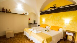 Una cama doble con ropa de cama amarilla y blanca, una mesita de noche de madera y un estante con objetos decorativos.