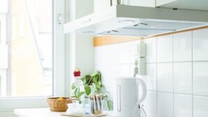 A modern kitchen with white cabinets, a stainless steel countertop, and a window with a floral curtain. - Duvan Hotell (Uppsala)