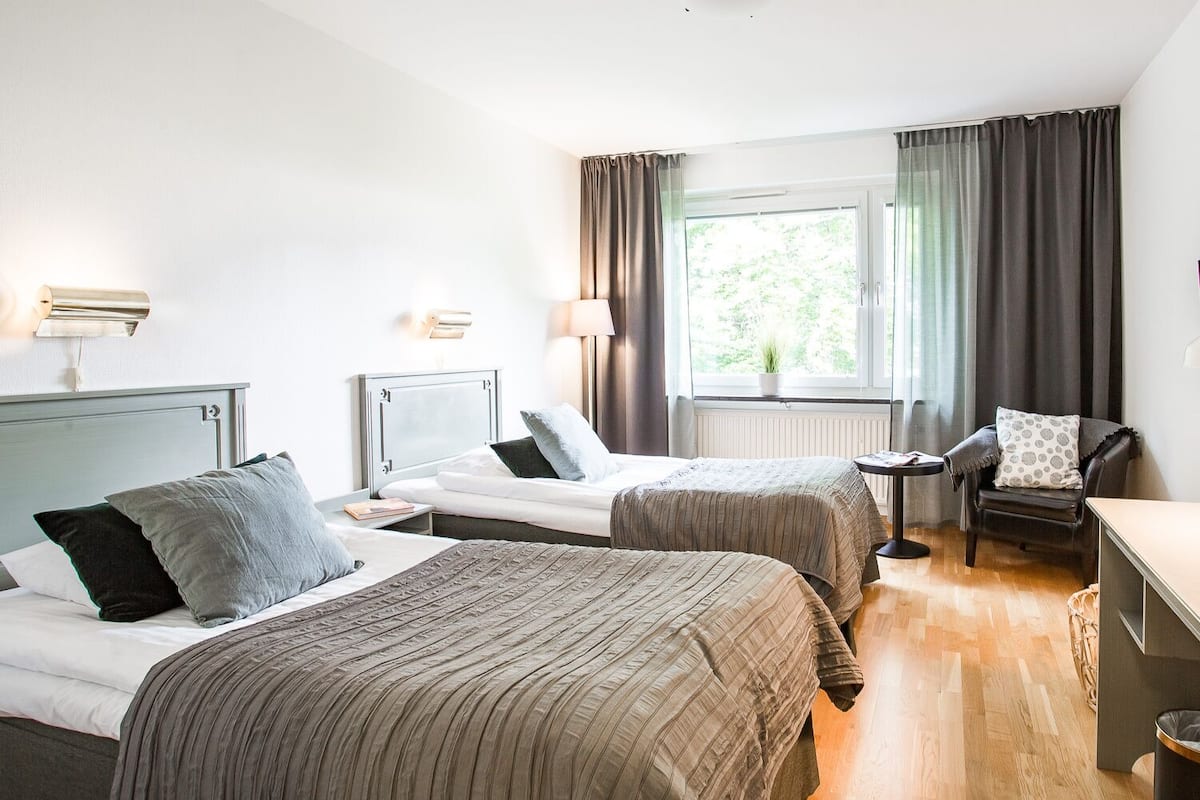 Ett hotellrum med två sängar, ett skrivbord, en stol och ett fönster med gardiner.