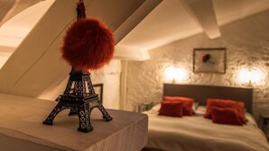 A model of the Eiffel Tower on a wooden shelf in a room with a bed and red pillows, a stone wall, and a framed picture on the wall. - Clos de la Colombe (Pouzols)