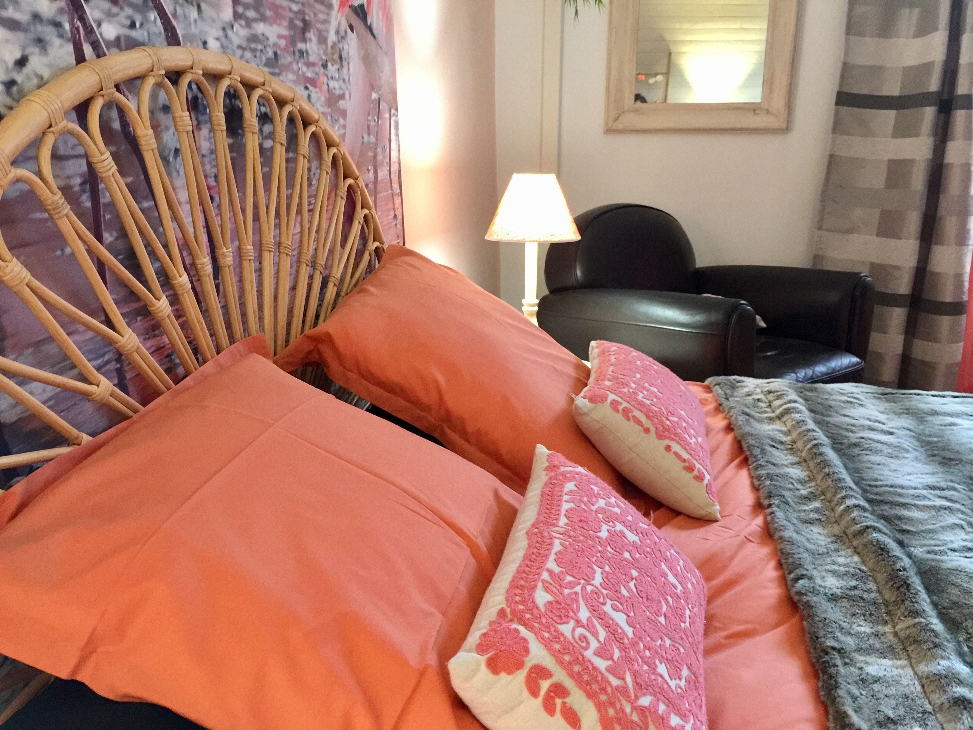 A room with a wicker chair, orange and pink pillows, a black leather chair, a lamp, and a checkered curtain.