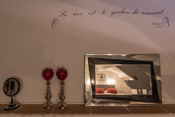 A wall with decorative items including a mirror, two red apples on stands, and a large mirror reflecting a room with a bed and pillows.
