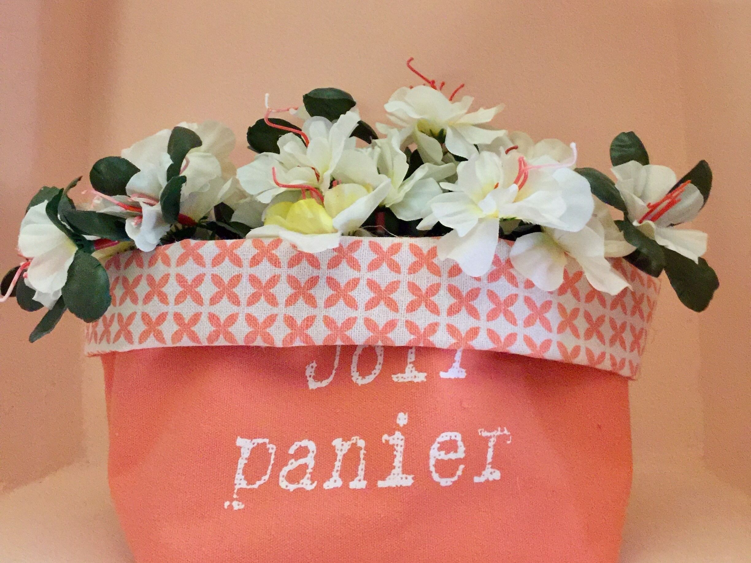 Une plante en pot, avec des fleurs blanches et des feuilles vertes, posée sur un sac en tissu orné d’un motif et sur lequel on peut lire le mot « panier ».