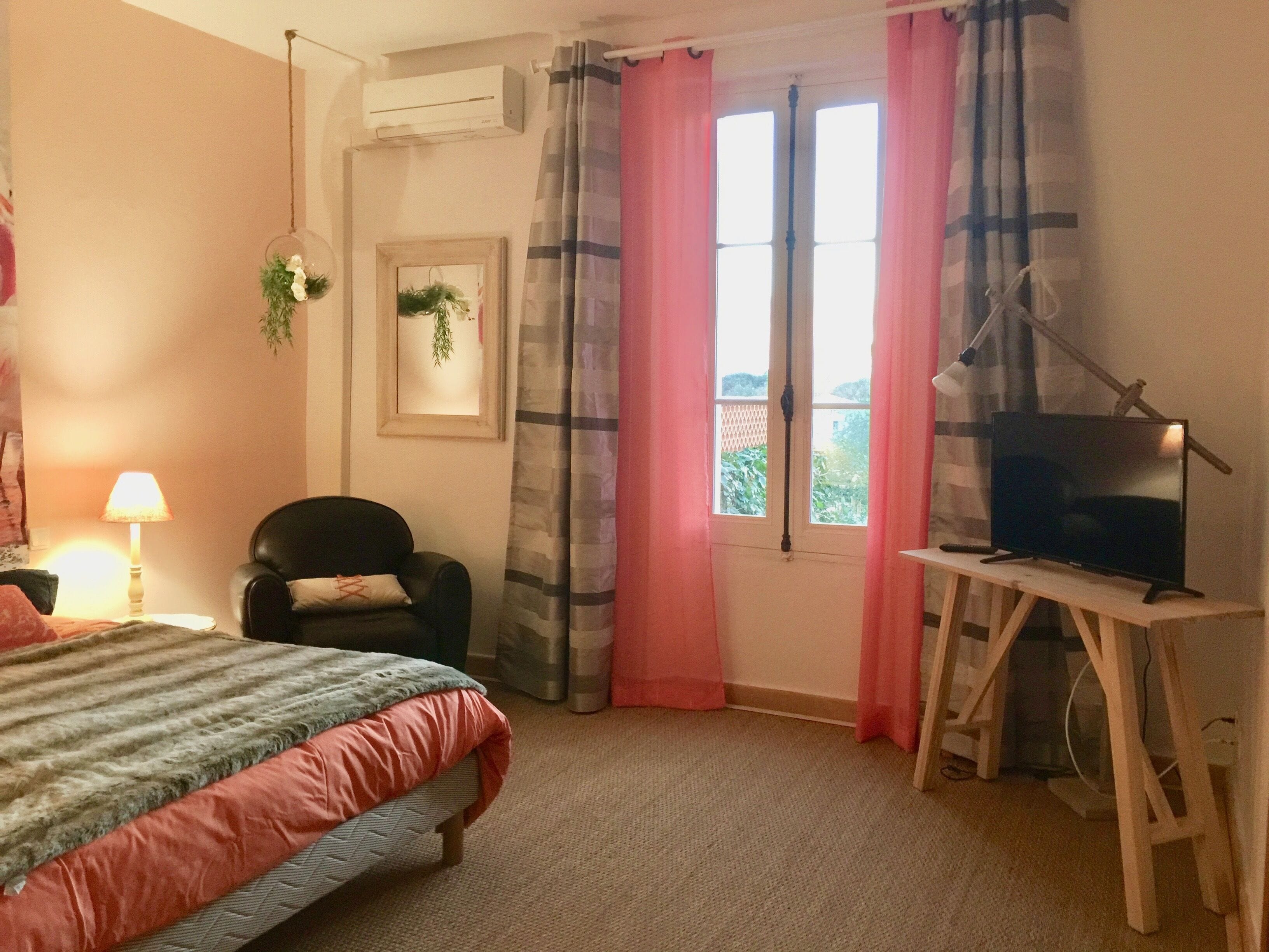 A bedroom with a bed, a chair, a desk with a TV, a window with curtains, and an air conditioning unit.