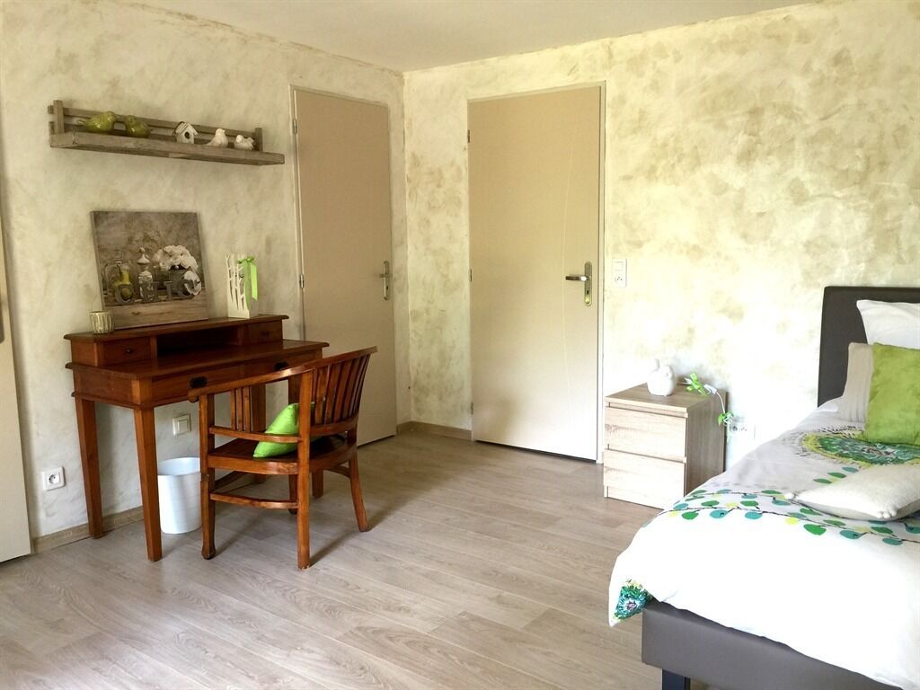 Een slaapkamer met een bed, een bureau en een stoel. Boven het bureau bevindt zich een plank en er is een deur op de achtergrond.