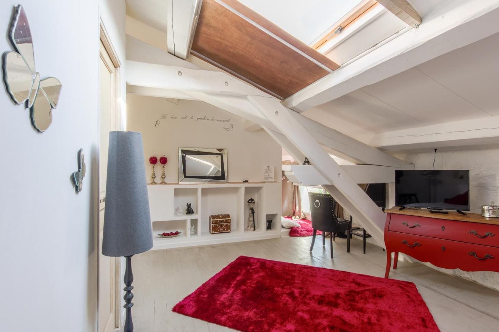 Une chambre à coucher aménagée dans un espace de type « loft », avec un puits de lumière, un tapis en velours rouge, une télévision et une commode rouge.