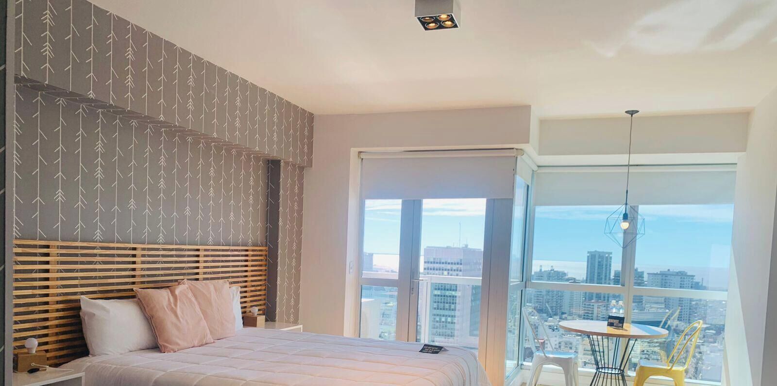 A modern bedroom with a large bed, a city view through a balcony, and a small table with chairs.