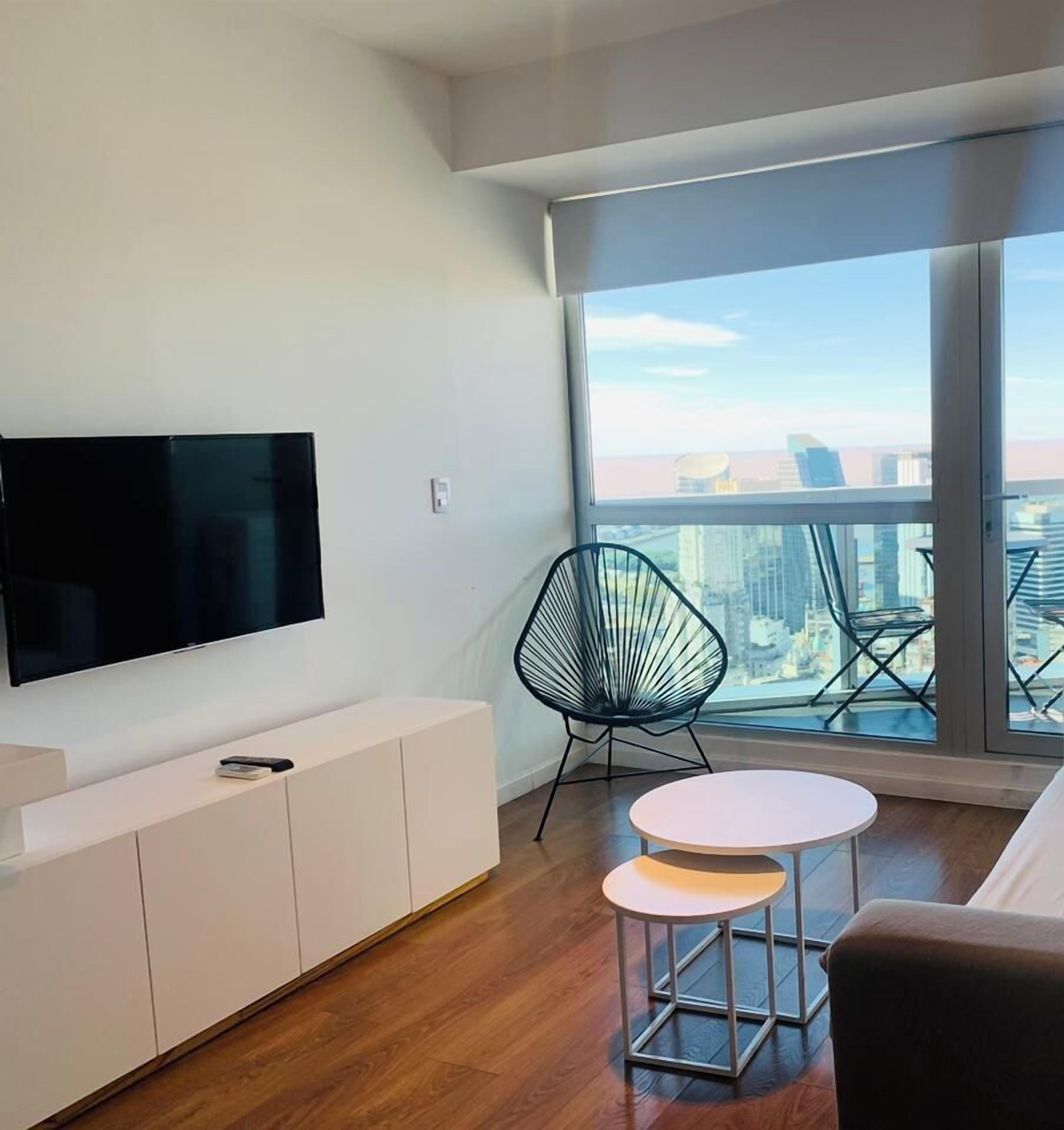 A modern living room with a flat-screen TV, a white console, two round tables, a black chair, and a large window with a city view.