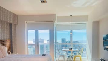 A bedroom with a bed, a table with chairs, and a city view through large windows.
