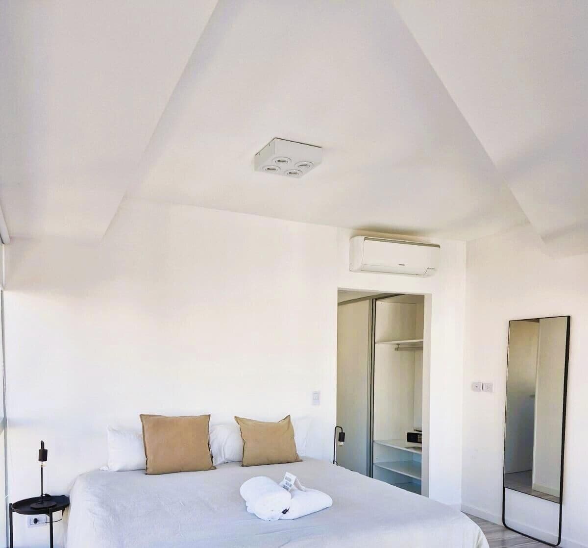 A hotel room with a bed, a bedside table, a mirror, and an air conditioning unit.
