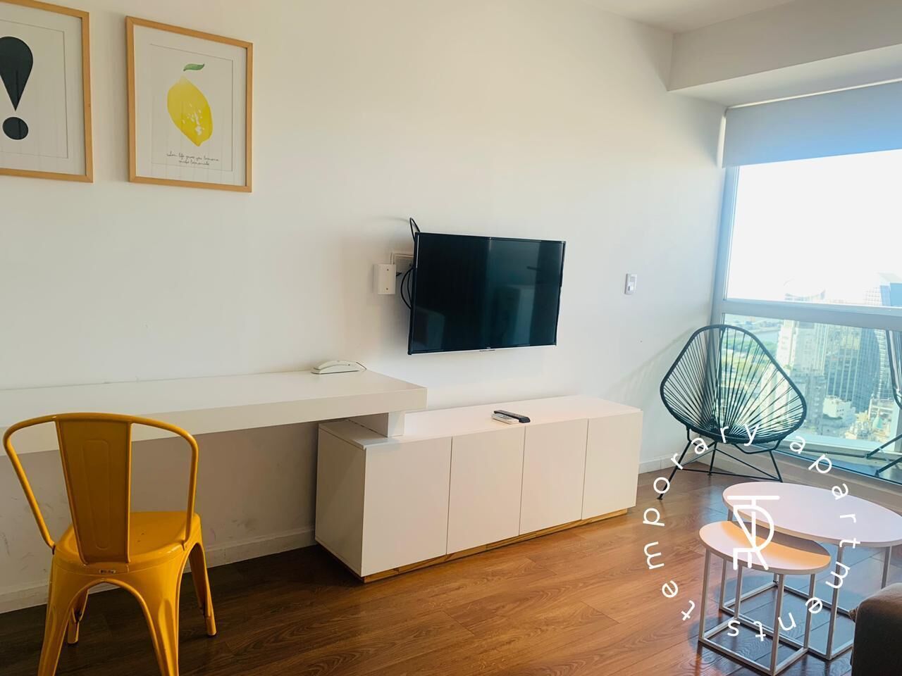 Un espacio habitable moderno con una televisión de pantalla plana montada en la pared, una consola blanca, una silla amarilla, una mesa redonda con sillas y vistas a la ciudad a través de amplios ventanales.