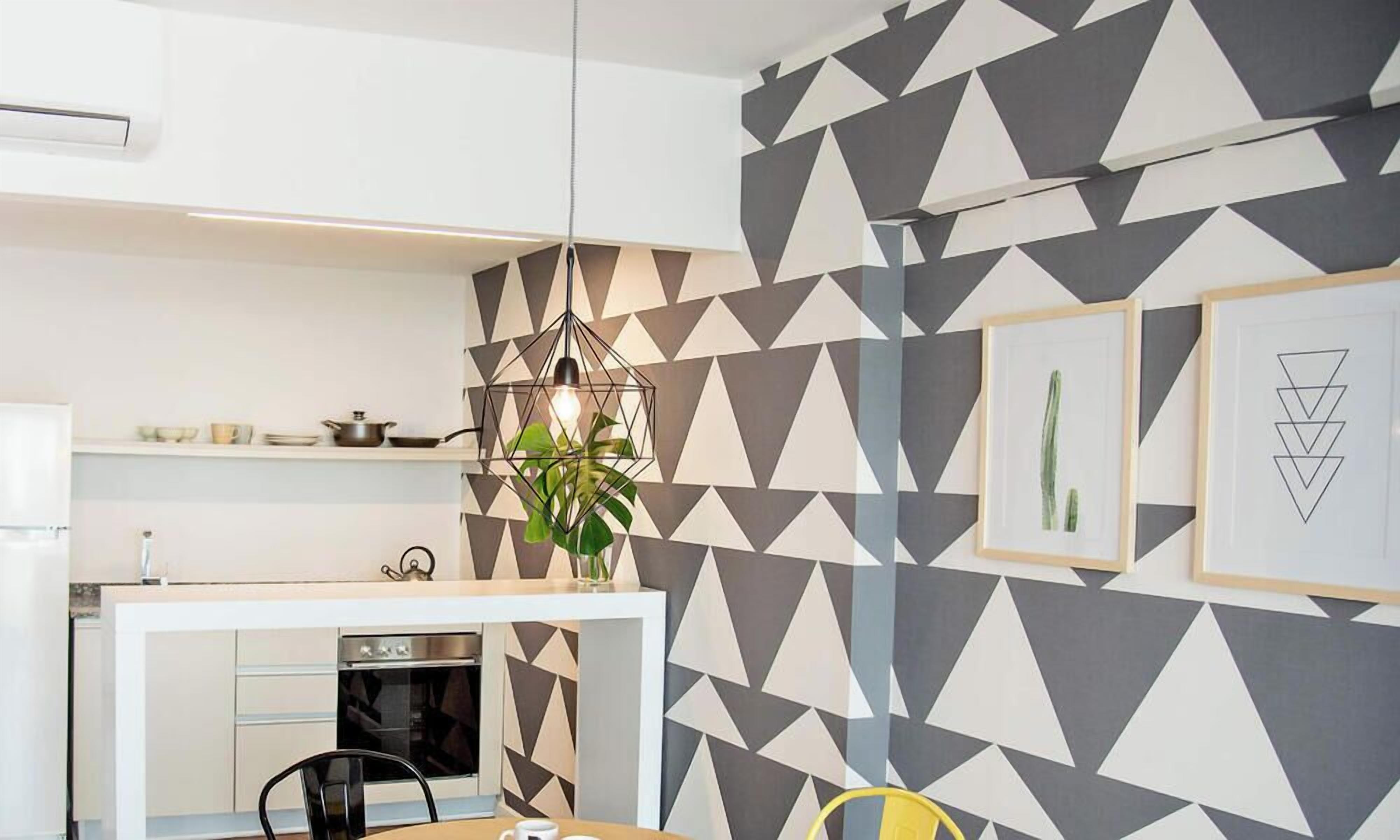 Una cocina moderna con un muro de estampado geométrico, una lámpara colgante y un área de comedor con una silla amarilla.