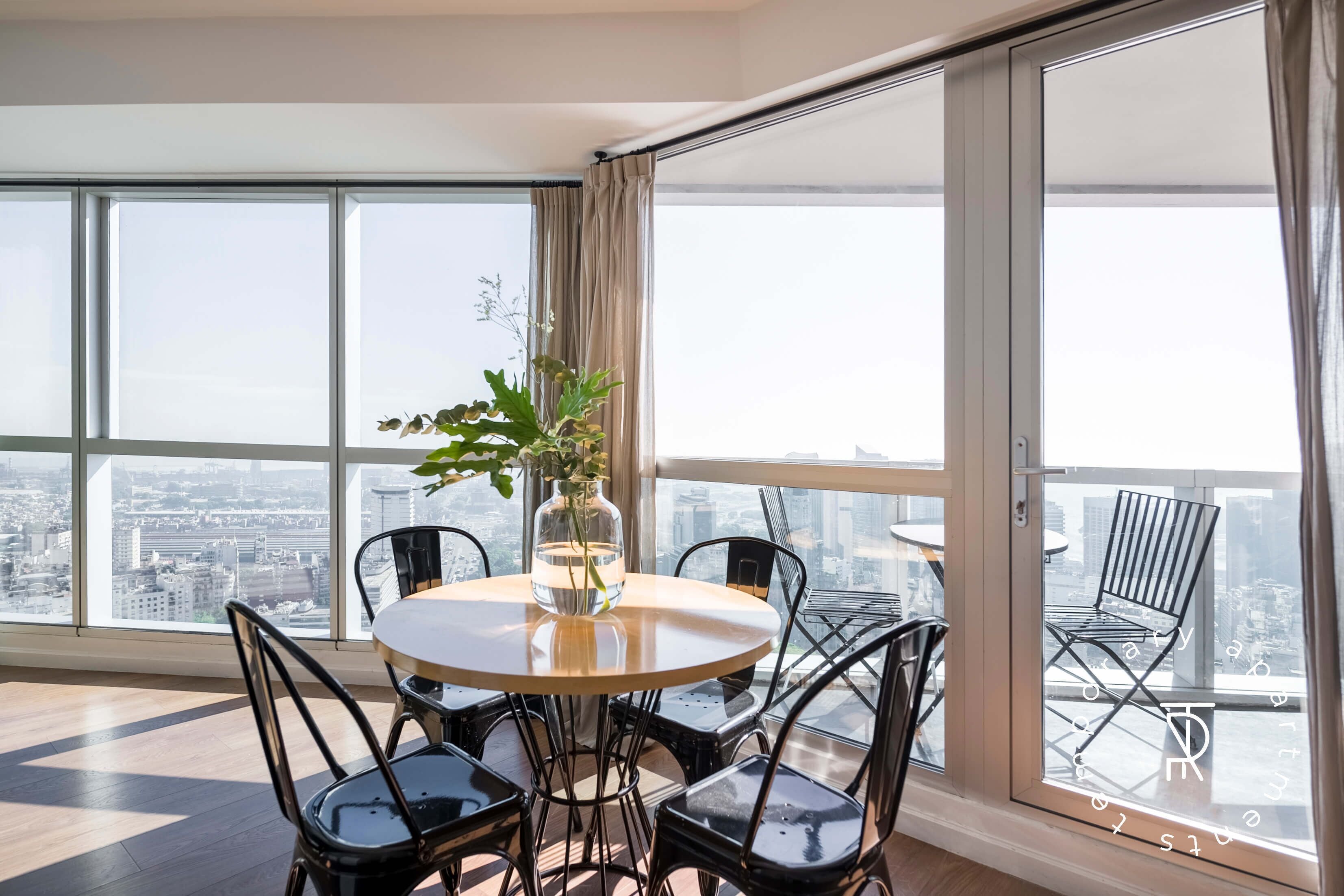 Un appartement situé dans un immeuble de grande hauteur, doté d’un grand balcon, d’une table de salle à manger ronde et de chaises en métal noir.