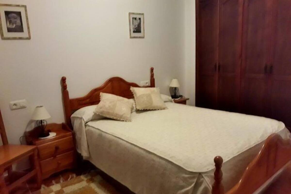 A neatly made bed with a wooden headboard, a bedside table with a lamp, and a checkered rug on the floor.