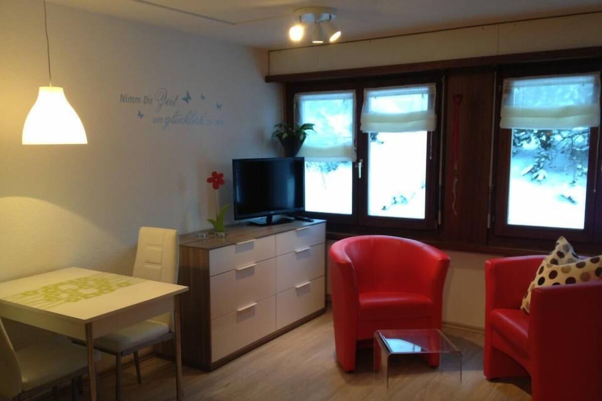 A modern living room with a dining area, a red armchair, a TV on a wooden cabinet, and large windows with blinds.