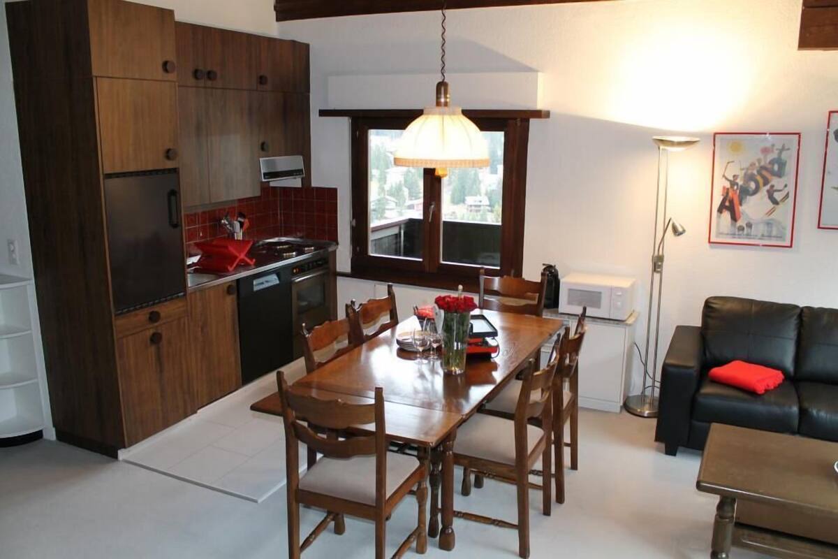 A modern kitchen with wooden cabinets, a dining table with chairs, and a black leather sofa in a living area.