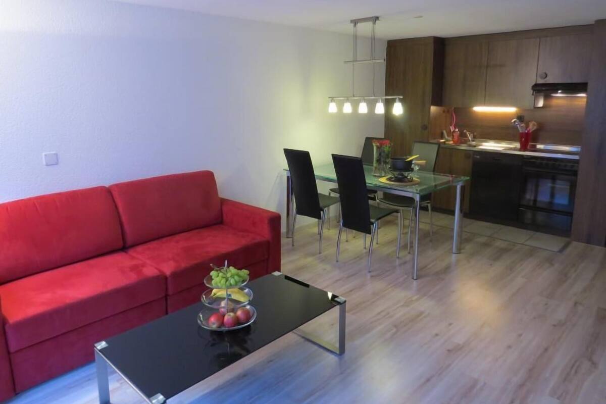 A modern living room with a red sofa, a glass dining table with chairs, and a kitchen area with wooden cabinets and a stove.