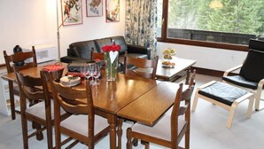 A dining area with a wooden table and chairs, a vase with red flowers, and a view of trees through a window.