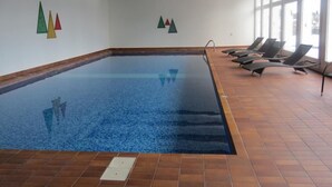 An indoor swimming pool with clear blue water, tiled floor, and sun loungers along the right side.