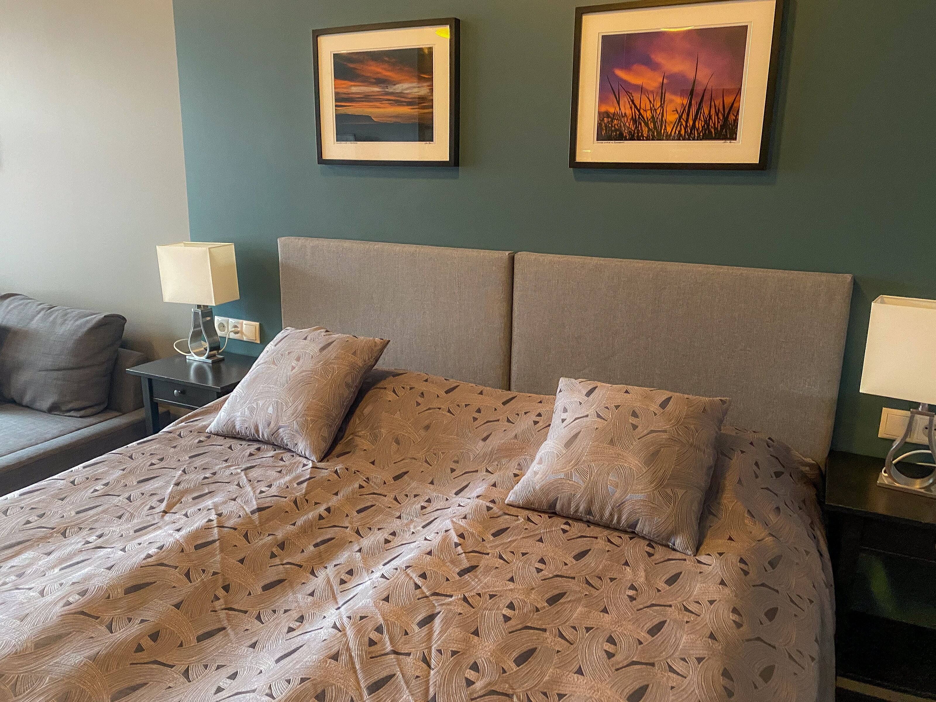 Ein ordentlich bezogenes Bett mit gemusterten Bettwäsche, einem grauen Kopfteil und zwei gerahmten Bildern an der Wand.
