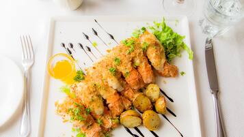 A white plate with breaded fish, potatoes, and greens, accompanied by a fork and knife on a white tablecloth.