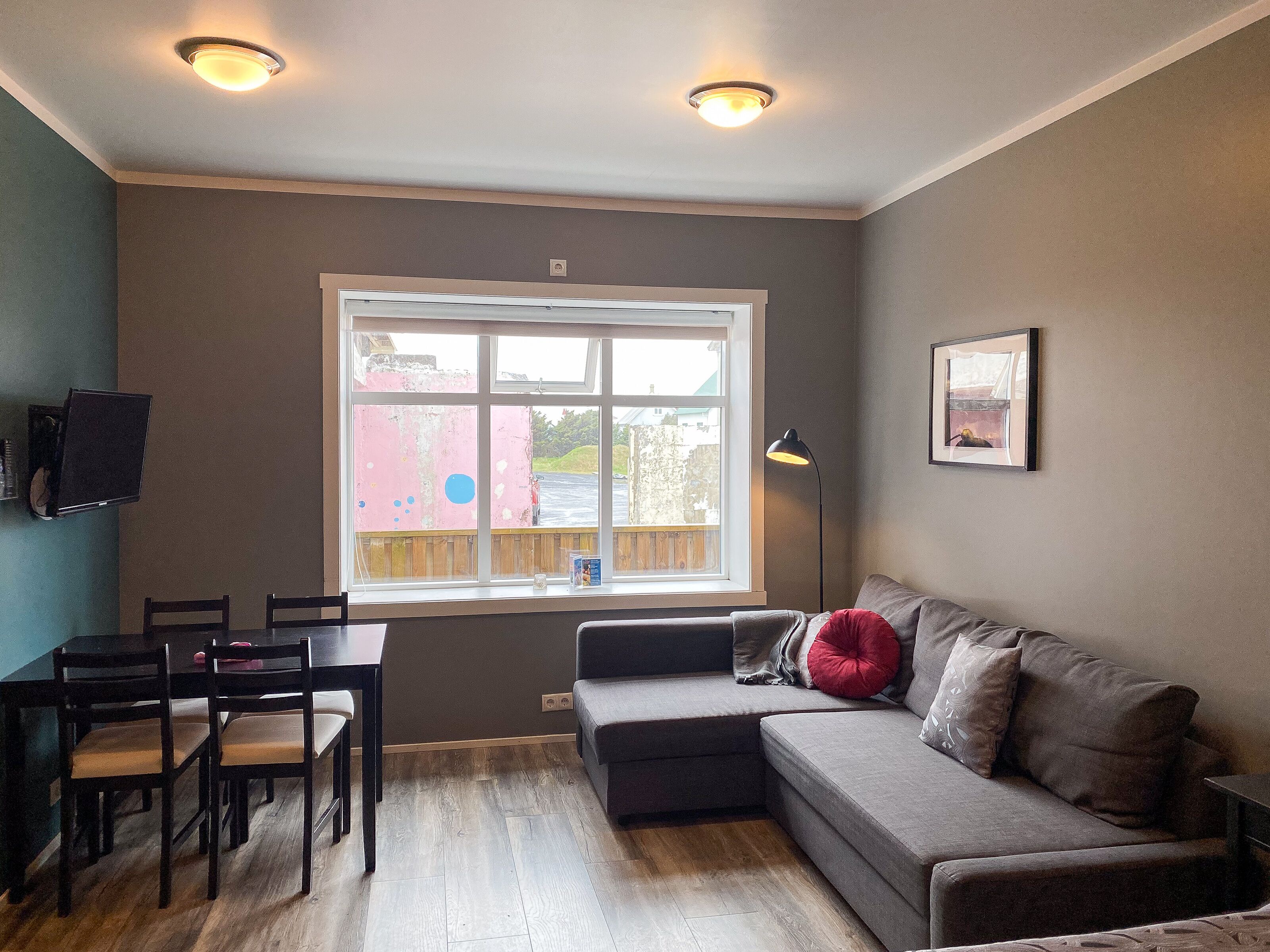 A living room with a gray sofa, a dining table with chairs, a window with a view of a pink building, and a ceiling light fixture.