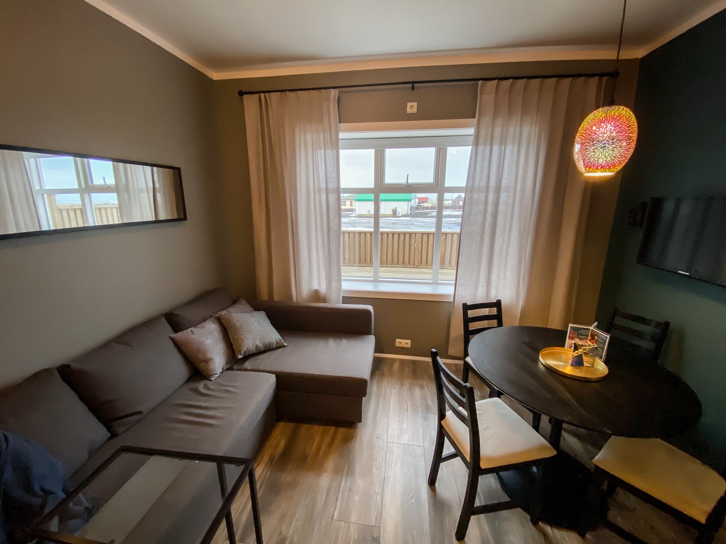 A modern living room with a sofa, a dining table with chairs, a large window with curtains, and a colorful hanging light fixture.