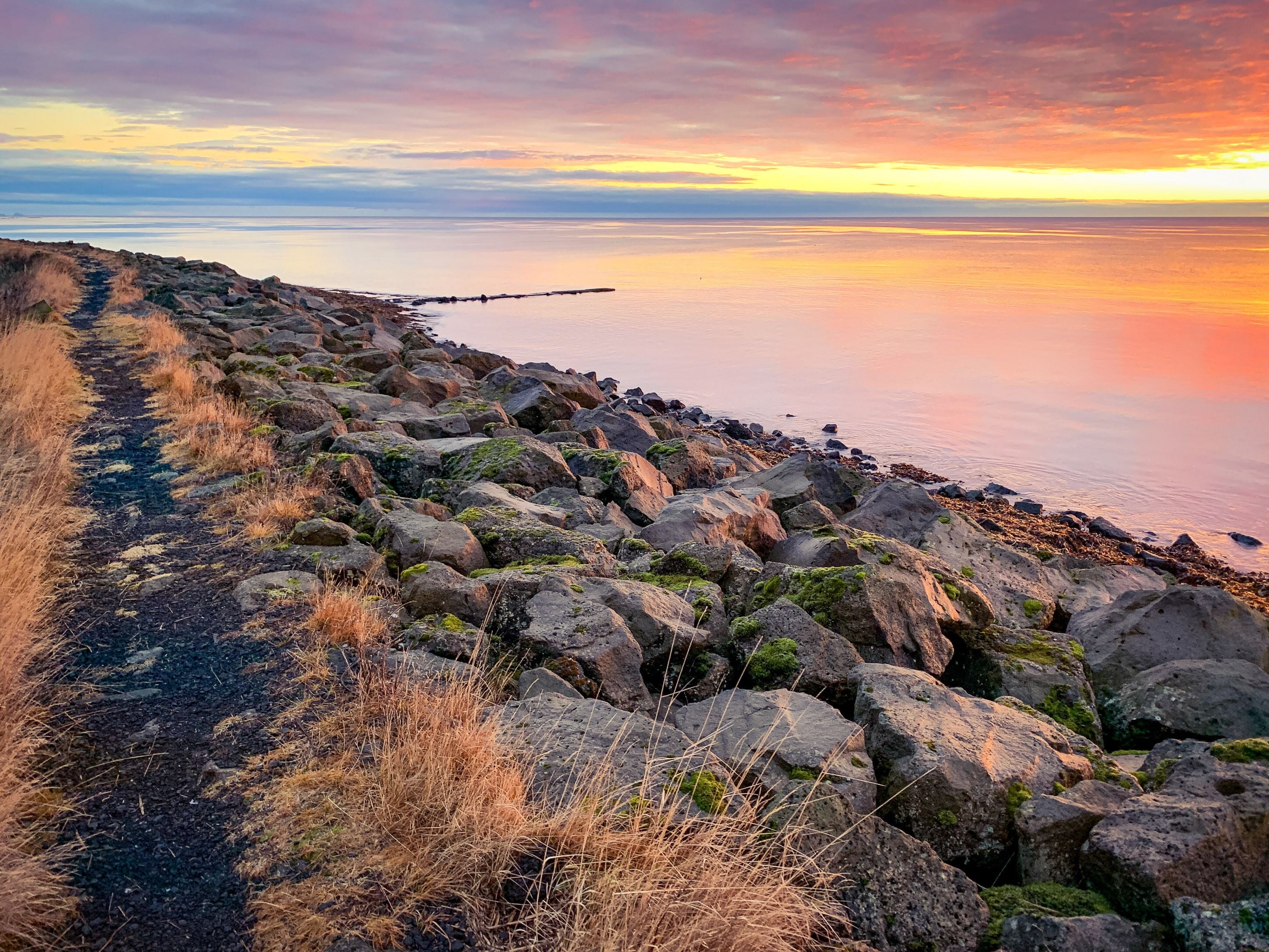 A rocky shoreline with a path leading to the water, a wooden pier extending into the sea, and a sunset in the background.