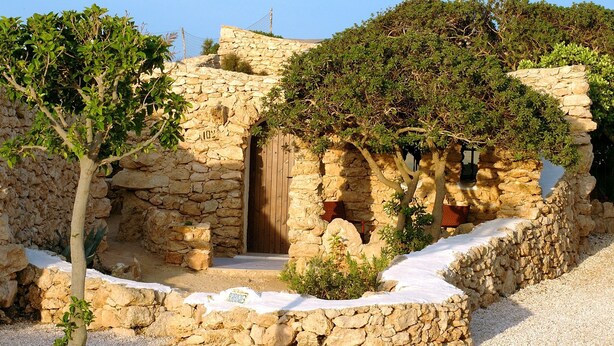 A stone building with a wooden door, surrounded by trees and a stone wall. - Albergo I Dammusi Di Borgo Cala Creta (Lampedusa)