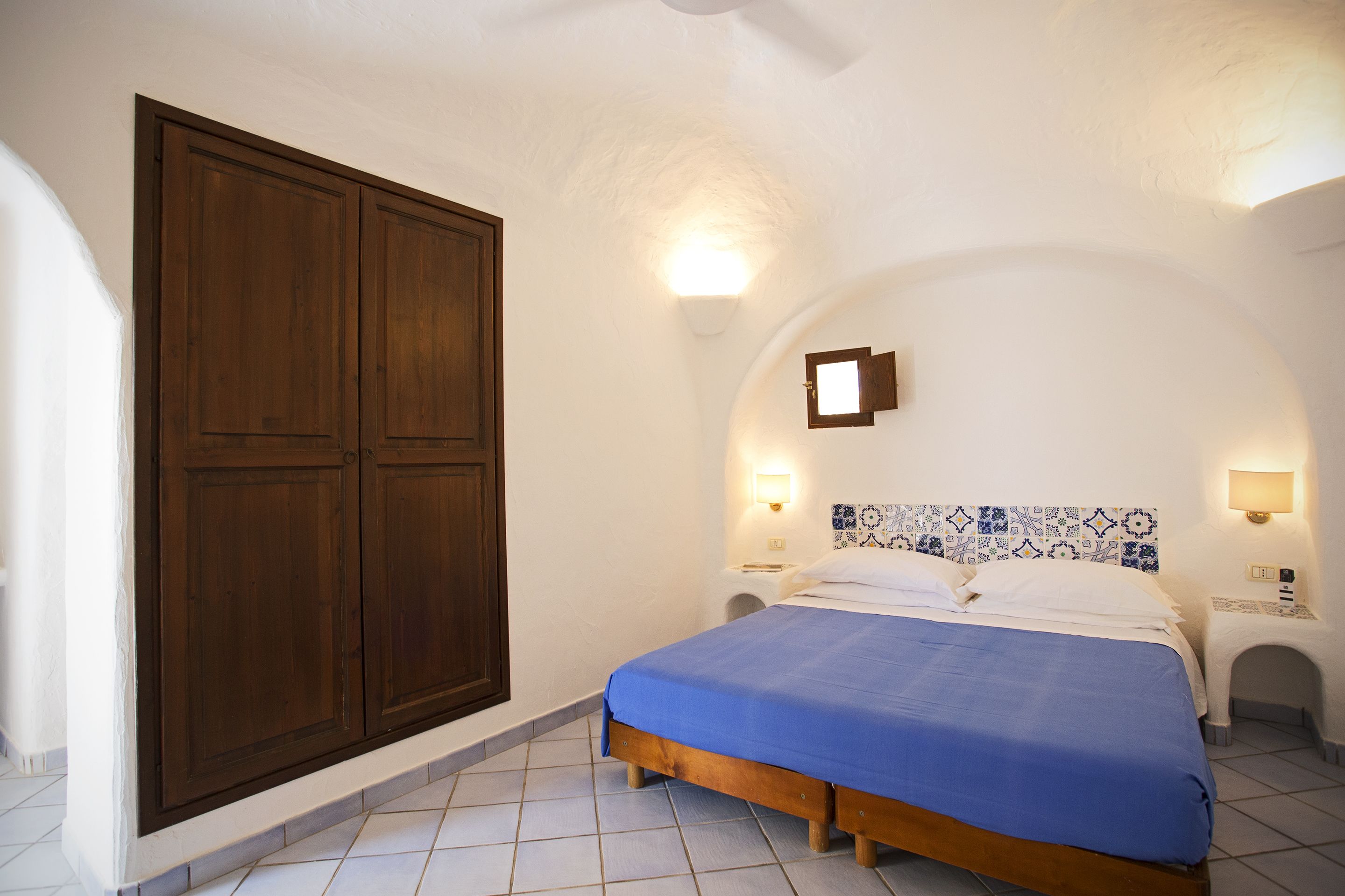 Une chambre avec une grande armoire en bois, un lit recouvert d’une courtepointe bleue et un sol carrelé.