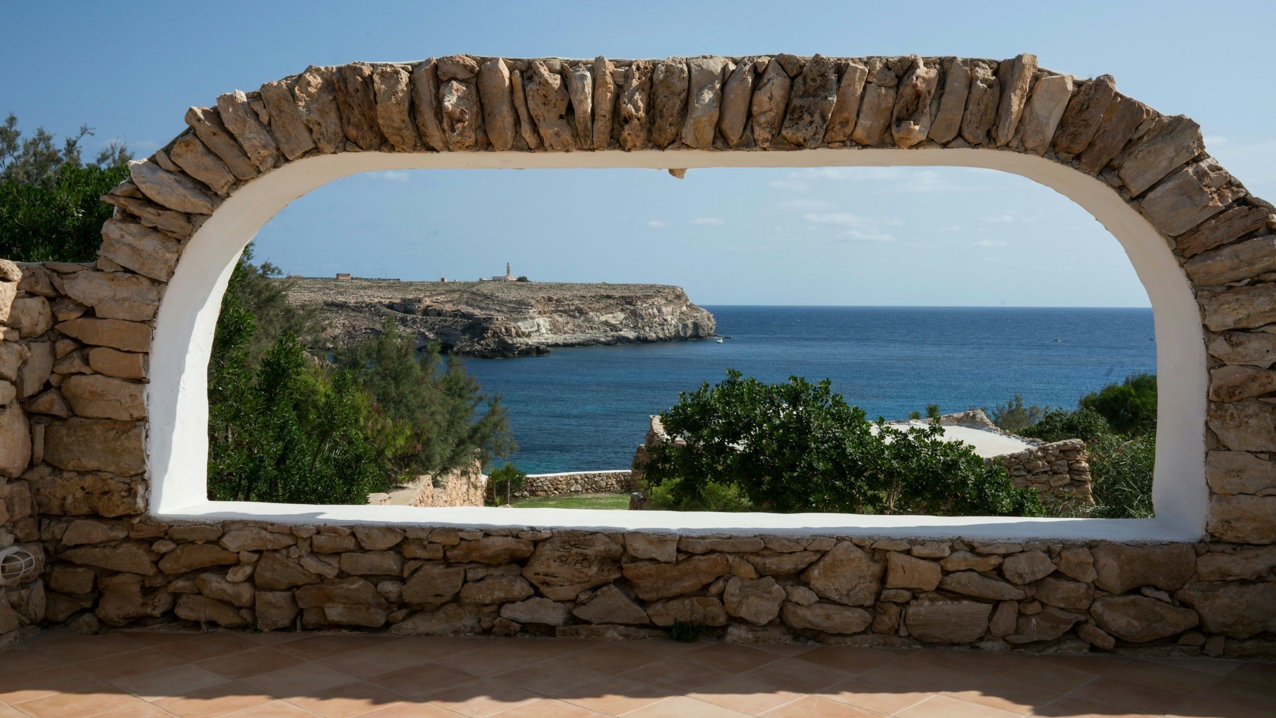 Ein steinrahmiges Fenster mit Blick auf eine Küstenlandschaft, einen Felsen und das Meer.