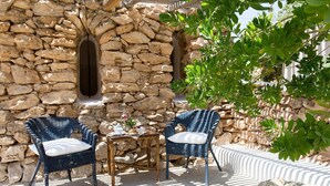 A stone wall with a window, two wicker chairs with cushions, a small round table with a teapot and cups, and a potted plant. - Albergo I Dammusi Di Borgo Cala Creta (Lampedusa)