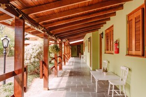 A covered outdoor corridor with wooden beams, a stone floor, and white chairs. - Pousada Villa Cantaloa Gramado (Gramado)