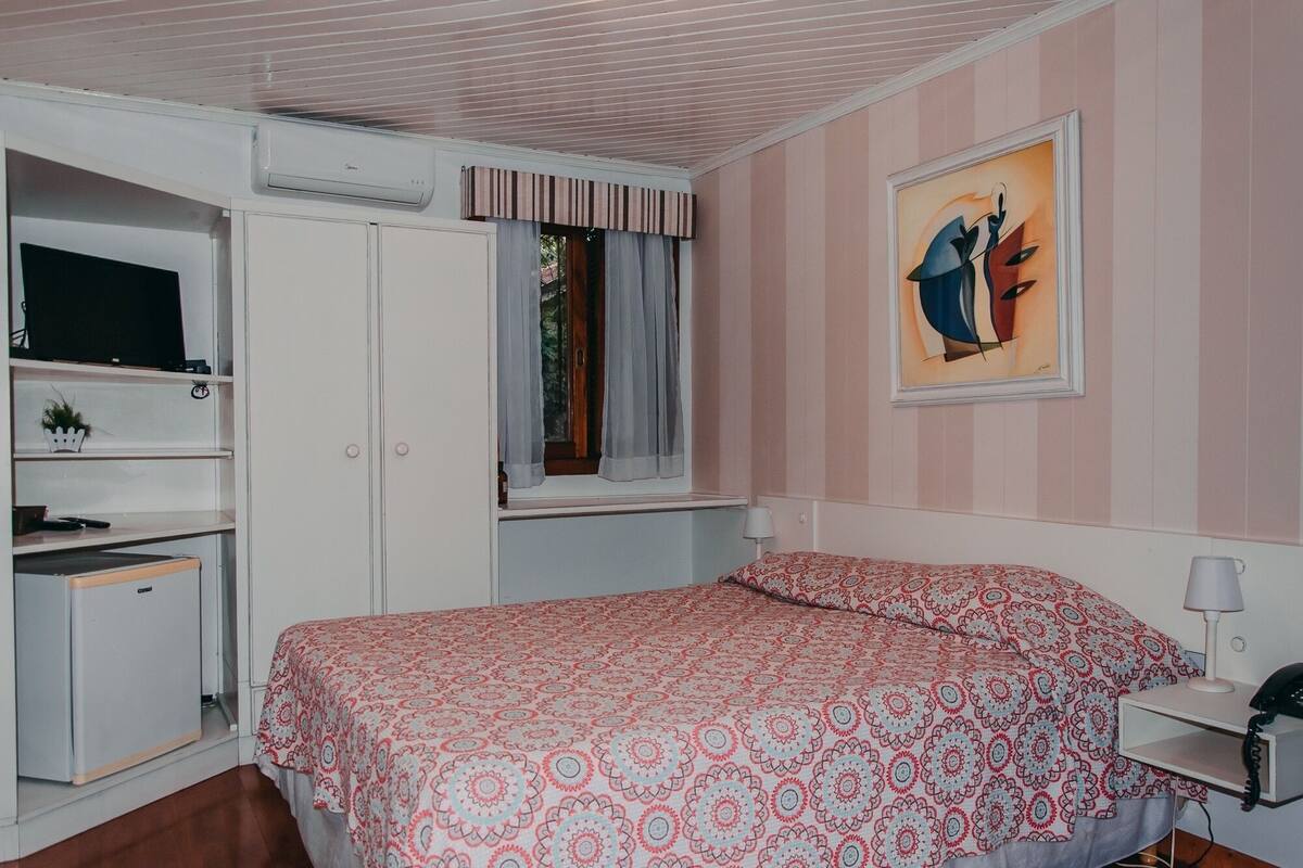 Un dormitori amb llit, escriptori, cadira, televisió i finestra amb cortines.